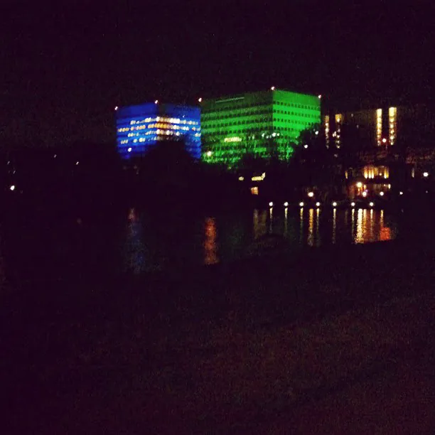 Office buildings lit in blue and green at night, reflected in the water