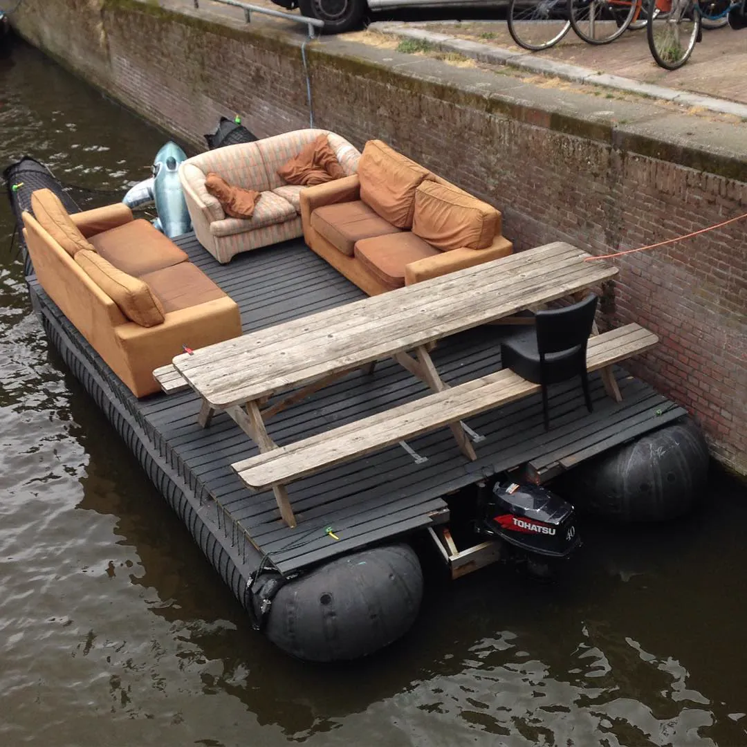 A floating living room: sofas and a picnic table on a raft moored in a canal