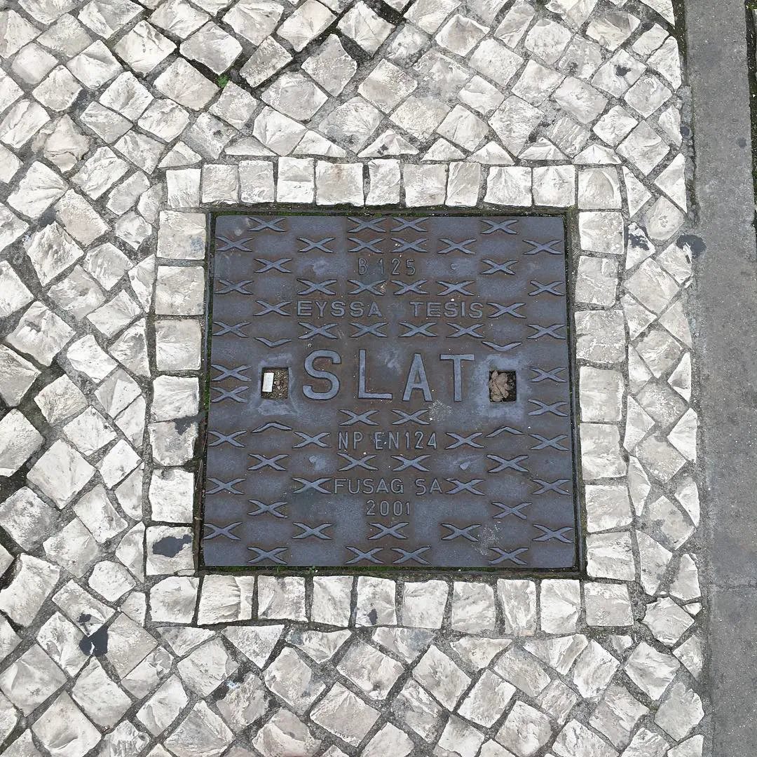 A square manhole cover framed by white Portuguese cobblestones, in Lisbon