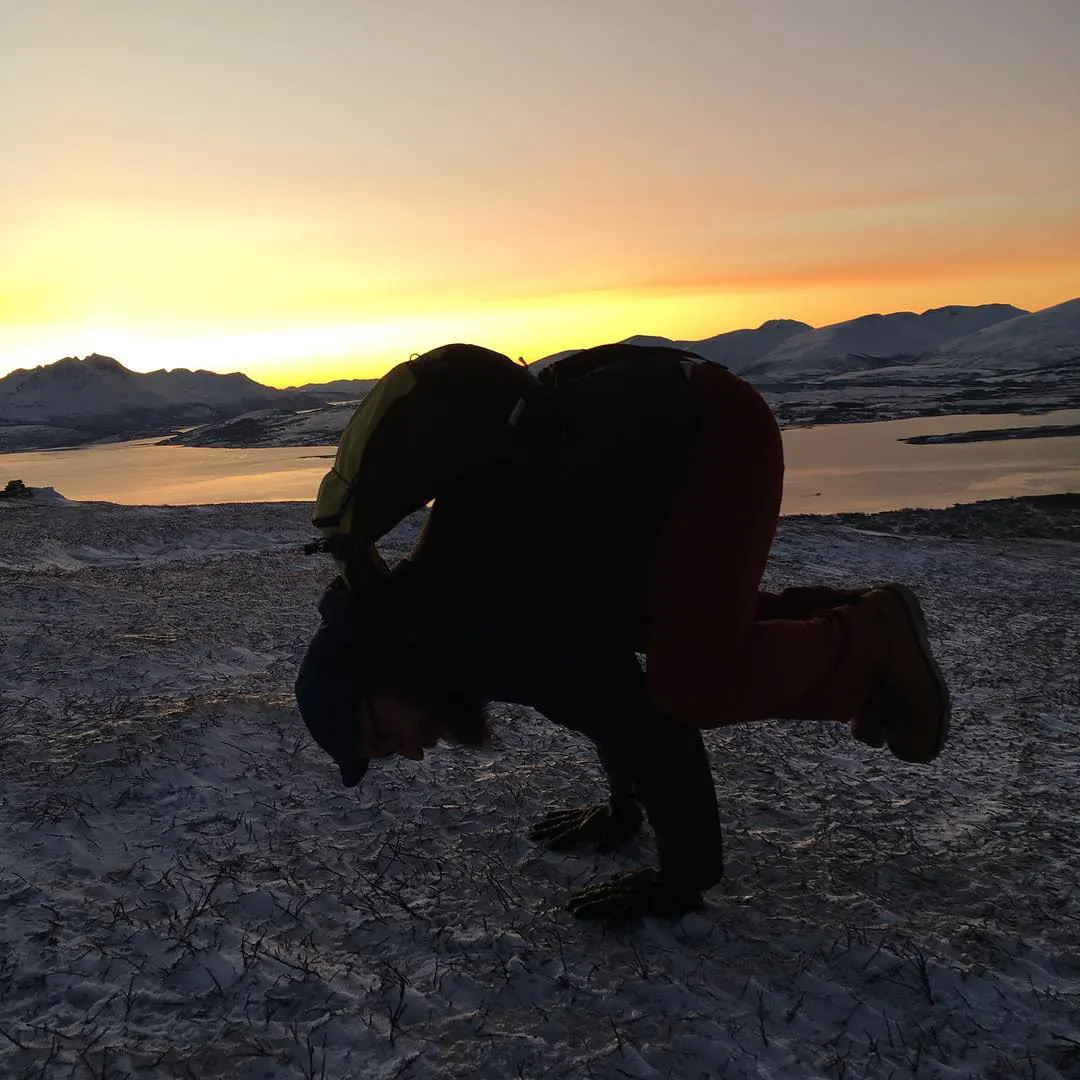Natan in crow pose in silhouette at sunset, with snowy hills and a fjord in Tromsø behind