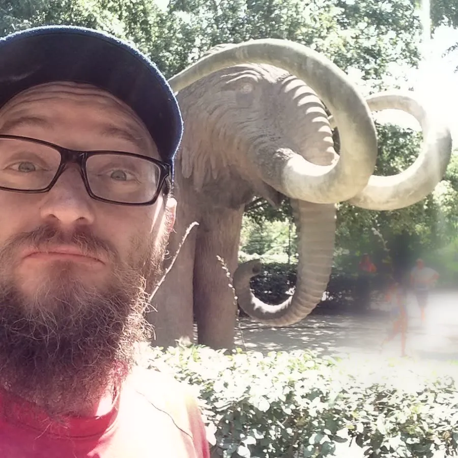 Natan's selfie in a cap, next to a life-size stone mammoth in a park